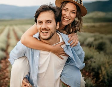 The image shows a smiling couple in a scenic outdoor setting. The man is giving the woman a piggyback ride and both are laughing. They are dressed casually, and the background features fields and mountains under a clear sky.